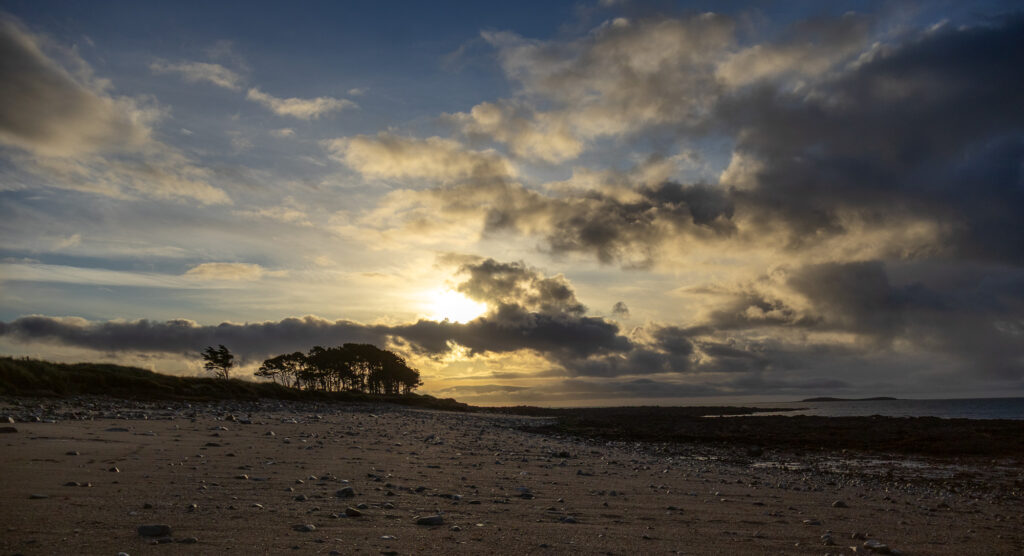 Une plage de sable parsemée de rochers au coucher du soleil, avec des arbres se découpant en silhouette à gauche, des nuages spectaculaires qui traversent le ciel, et le soleil partiellement caché derrière les nuages près de l'horizon.