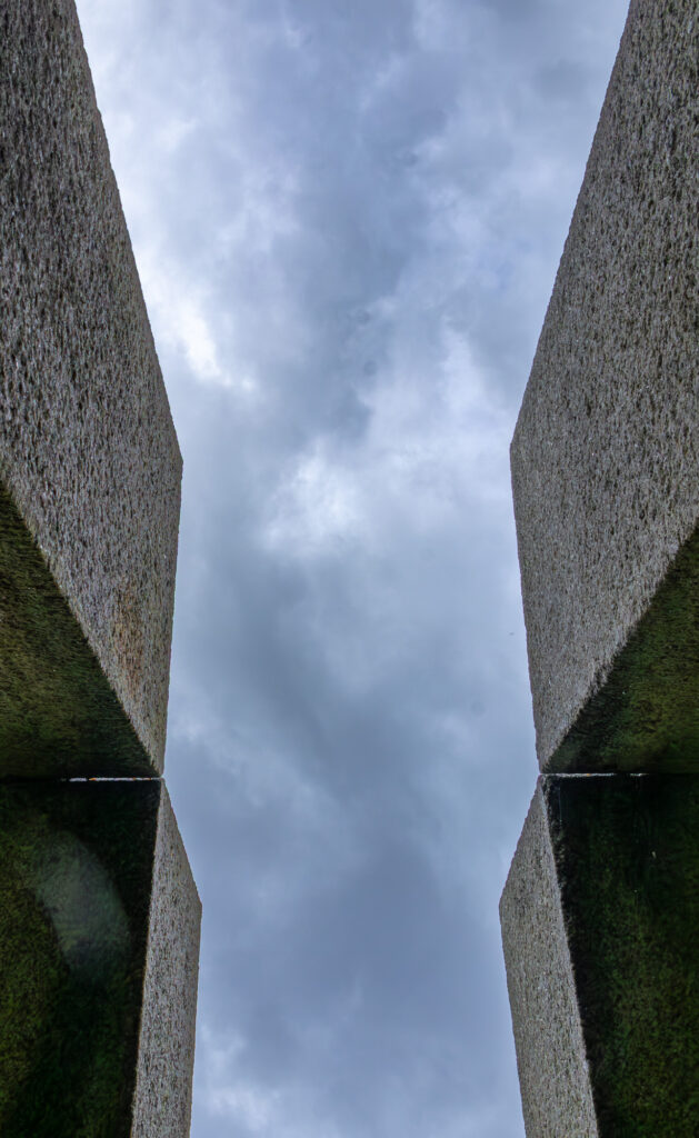 Vue vers le haut entre deux hauts murs en béton aux surfaces texturées, formant un étroit passage qui encadre un ciel nuageux et couvert au-dessus.