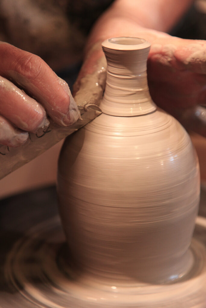 Gros plan sur des mains qui façonnent de l'argile humide sur un tour de potier, pour créer un vase à col étroit. Une main lisse la surface tandis que l'autre guide l'argile ; toutes deux sont recouvertes d'argile humide.