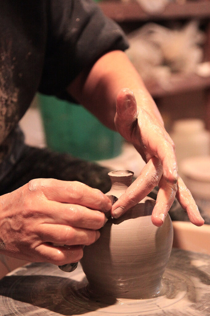 Une personne façonne un petit vase en argile sur un tour de potier, en utilisant ses mains pour lisser et modeler la surface. Ses mains sont recouvertes d'argile humide, ce qui met en valeur le processus de fabrication de la poterie.