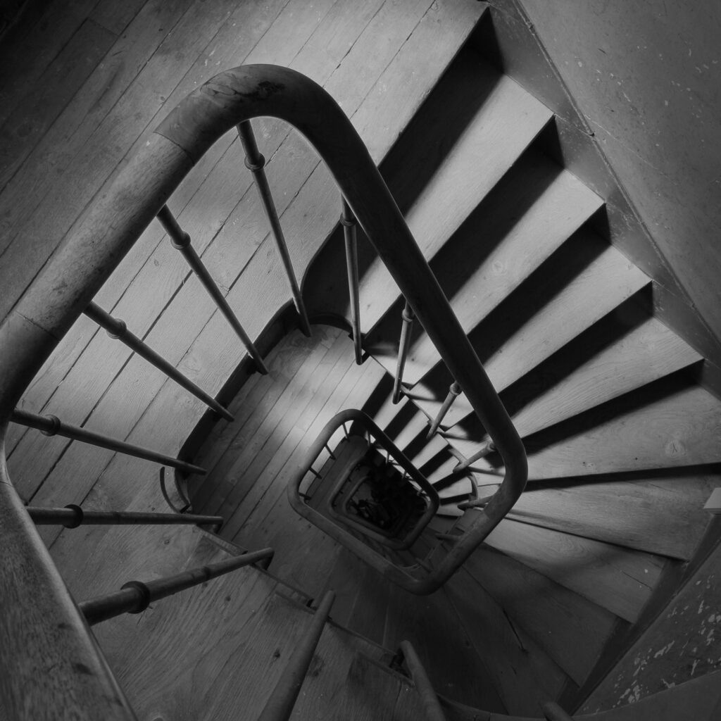 Photo en noir et blanc d'un escalier en colimaçon vu d'en haut, montrant des marches en bois et une rampe incurvée avec des balustres métalliques, formant un motif géométrique qui s'enroule vers le bas.
