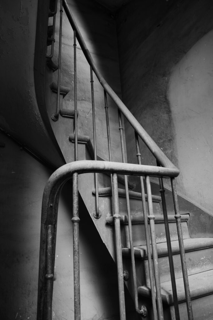 Photo en noir et blanc d'un vieil escalier étroit et sinueux, doté de rampes métalliques et de marches usées, qui monte le long d'un mur texturé dans un espace faiblement éclairé.