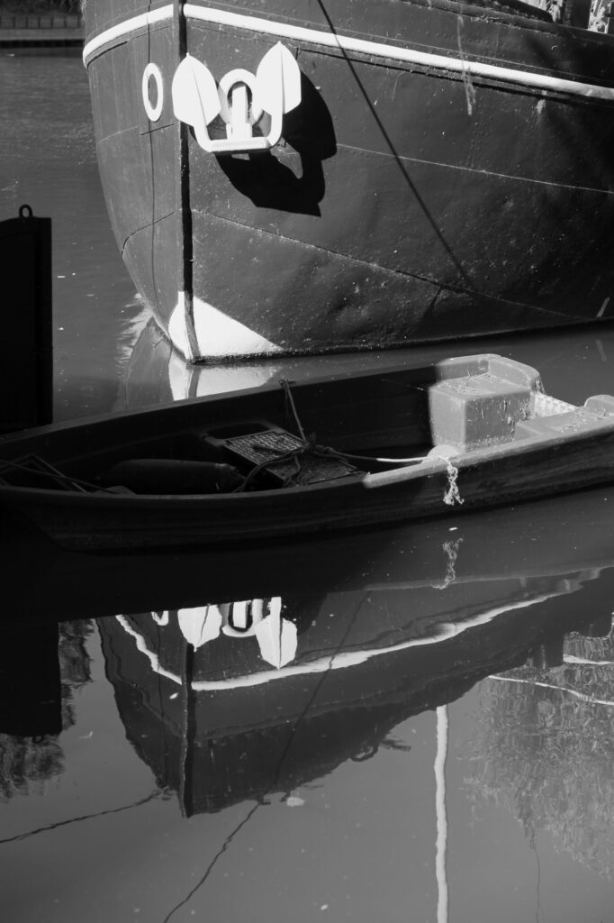 Photo en noir et blanc montrant la proue et l'ancre d'un grand bateau se reflétant dans une eau calme, avec une petite barque flottant au premier plan, partiellement dans l'ombre.