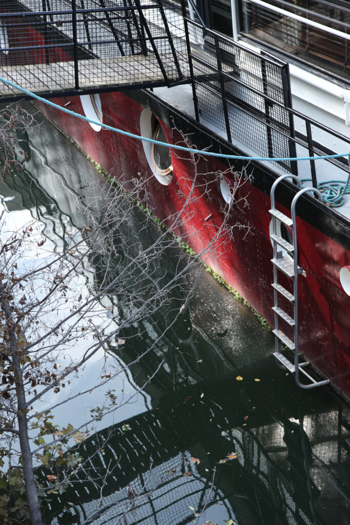 Une échelle métallique descend depuis un quai vers l'eau sombre, à côté d'un bateau rouge aux hublots ronds ; on aperçoit des branches nues et des reflets à la surface de l'eau.
