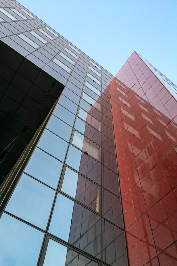 Vue en contre-plongée de deux gratte-ciel modernes aux façades de verre et de pierre rouge, se reflétant l'un dans l'autre, sur fond de ciel bleu limpide.