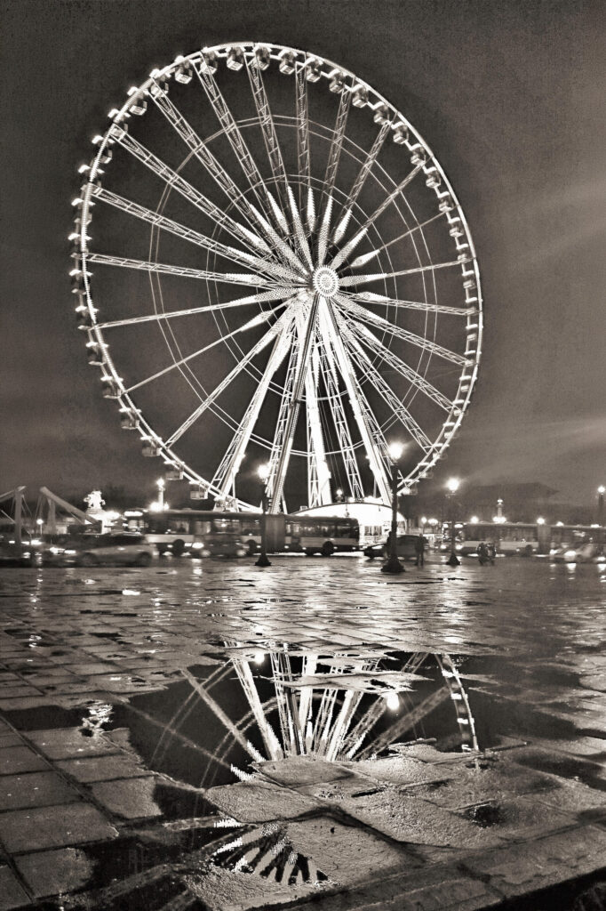 Une grande roue brillamment éclairée la nuit se reflète dans une grande flaque d'eau sur la chaussée mouillée. La scène dégage un effet spectaculaire, presque monochrome, avec la grande roue qui domine l'image et les lumières de la ville visibles en arrière-plan.