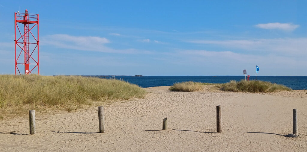 Une plage de sable avec, au premier plan, de hautes herbes et des poteaux en bois ; à gauche, une tour d'observation métallique rouge ; et, à l'arrière-plan, la mer bleue sous un ciel dégagé.