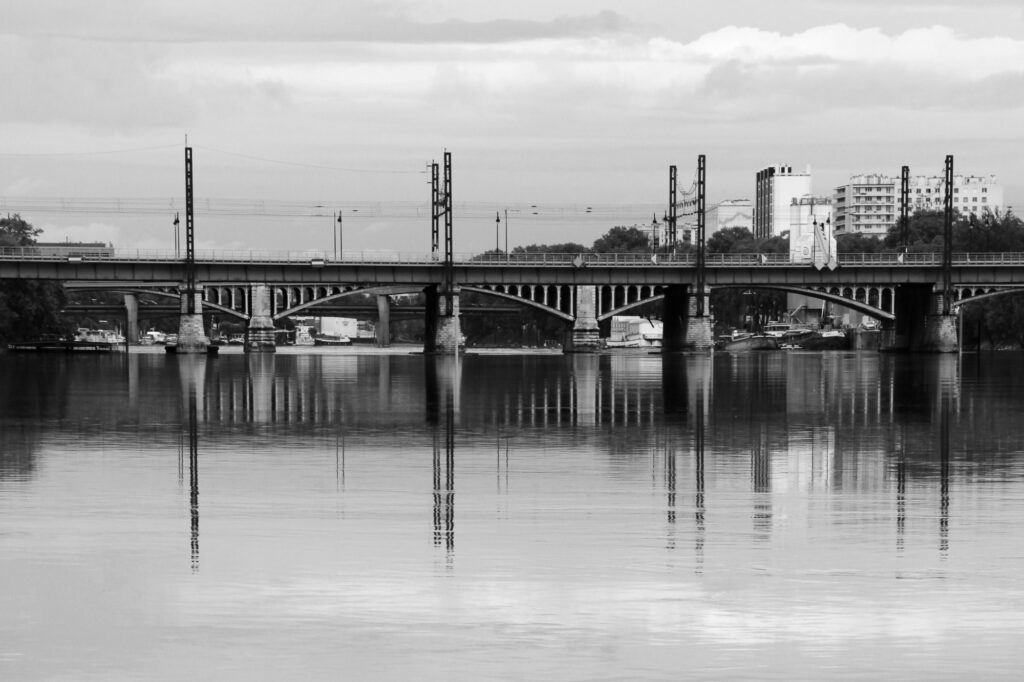 Une photo en noir et blanc d'un pont enjambant une rivière calme, dont le reflet se distingue dans l'eau. On aperçoit en arrière-plan des bâtiments, des arbres et un ciel nuageux.