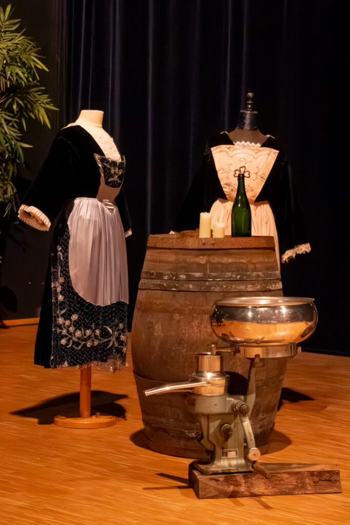 Deux mannequins vêtus de costumes traditionnels se tiennent à côté d'un tonneau en bois sur lequel sont posés des bougies, une bouteille de vin et une vieille machine en métal, le tout sur un plancher en bois. On aperçoit une plante verte à gauche.