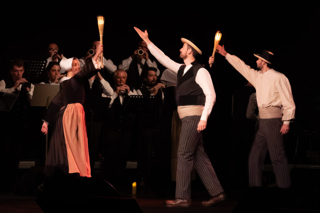 Trois artistes en costumes traditionnels, tenant des torches et dansant sur scène, se trouvent devant un orchestre assis. L'arrière-plan est sombre, ce qui met en valeur les danseurs et les musiciens.
