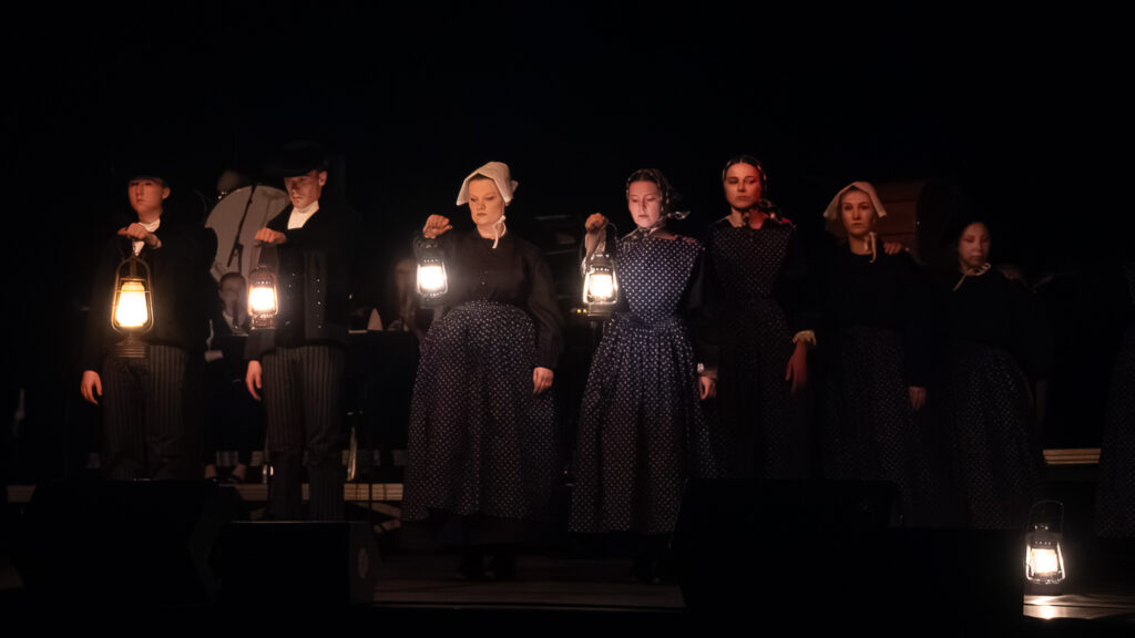Sept personnes vêtues de vêtements sombres et d'époque se tiennent en rang sur une scène faiblement éclairée, chacune tenant une lanterne allumée, créant ainsi une atmosphère sombre et dramatique sur fond noir.