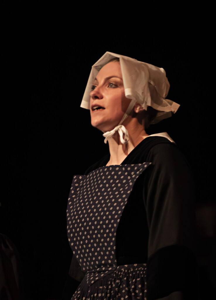 Une femme portant un bonnet blanc et une robe sombre à motifs, dotée d'un plastron, se tient debout devant un fond sombre, le regard légèrement levé vers le haut, l'air grave, évoquant un décor historique ou théâtral.
