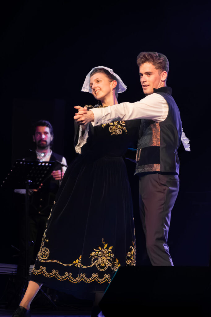 Deux jeunes gens vêtus de costumes folkloriques traditionnels dansent ensemble sur scène, tout en souriant. La jeune femme porte une robe sombre ornée de broderies dorées et un foulard blanc, tandis que le jeune homme porte un gilet. Un musicien joue en arrière-plan.
