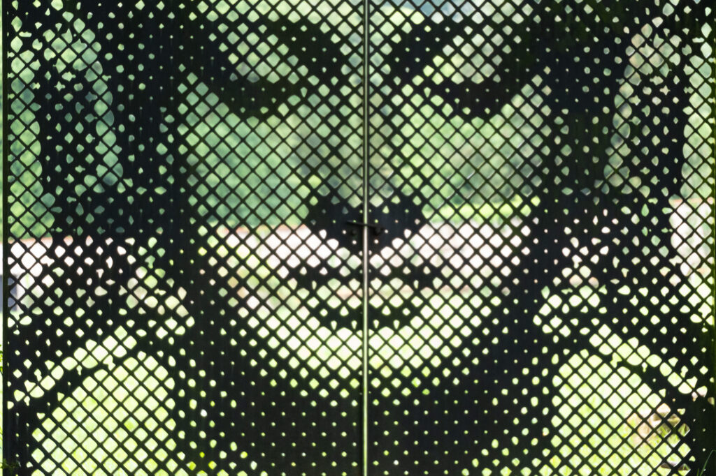 Panneau métallique perforé représentant, sous forme abstraite et pixélisée, un visage serein, peut-être celui de Bouddha, dans des tons verts et noirs, avec des rayons de soleil tamisés qui filtrent à travers les perforations.