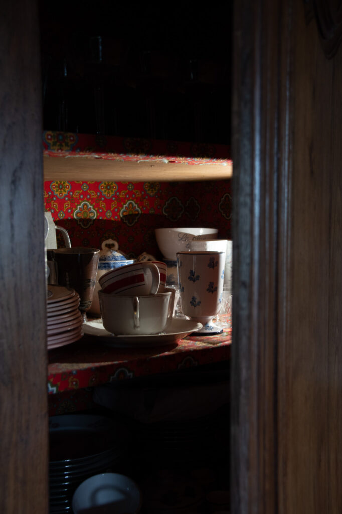 Une armoire en bois faiblement éclairée, dotée d'étagères à motifs, contient des assiettes, des mugs et des tasses empilés, dont certains arborent des motifs floraux. La lumière provenant de la gauche met en valeur la vaisselle, tandis que les étagères du bas restent dans l'ombre.
