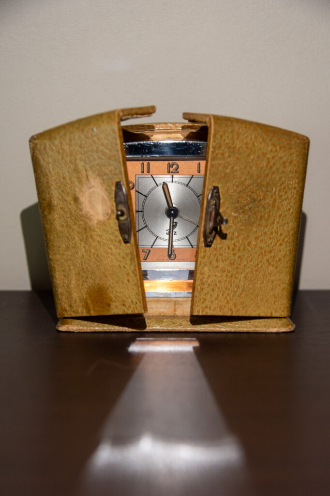 Une horloge ancienne, dans un boîtier usé de couleur brun jaunâtre, repose sur une surface sombre, son couvercle étant partiellement ouvert. La lumière éclaire l'horloge, projetant une ombre triangulaire sur la surface en dessous.
