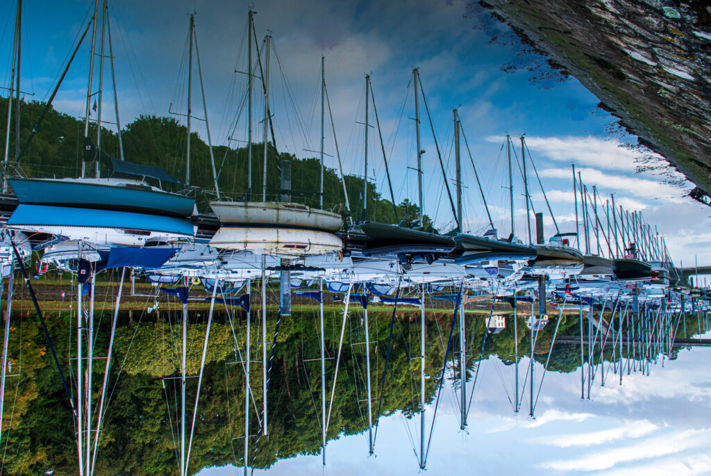 Plusieurs bateaux sont amarrés dans un port de plaisance ; leurs mâts se reflètent dans l'eau calme, créant un effet de miroir. On aperçoit à l'arrière-plan des arbres d'un vert luxuriant et un mur de pierre.