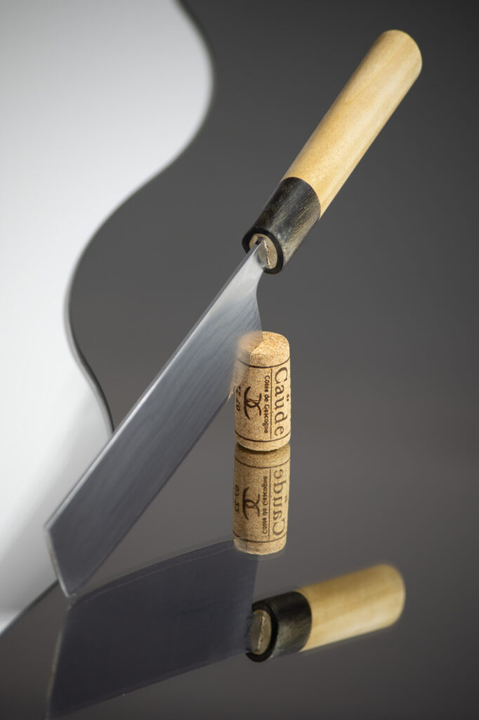 Un couteau de cuisine au manche en bois est posé en équilibre sur sa lame, au sommet de deux bouchons de vin empilés, le reflet des objets se voyant sur la surface brillante en dessous.