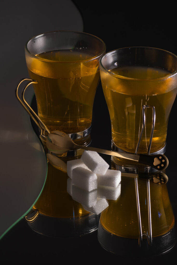 Deux verres à thé aux anses dorées reposent sur une surface noire brillante, à côté de quatre morceaux de sucre et de deux cuillères décorées. Cette composition crée de forts reflets et dégage une atmosphère à la fois mystérieuse et élégante.