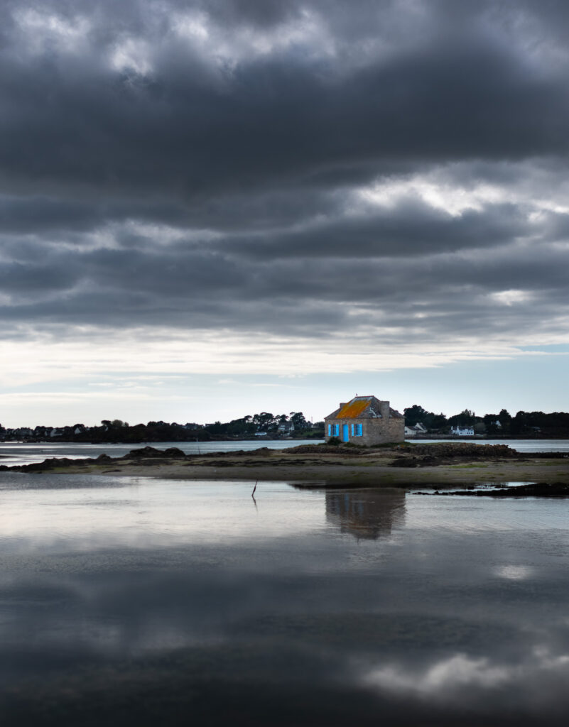 Une petite maison en pierre, au toit jaune et aux volets bleus, se dresse sur une pelouse au bord d'un plan d'eau tranquille, sous un ciel nuageux et spectaculaire, son reflet se dessinant à la surface de l'eau.
