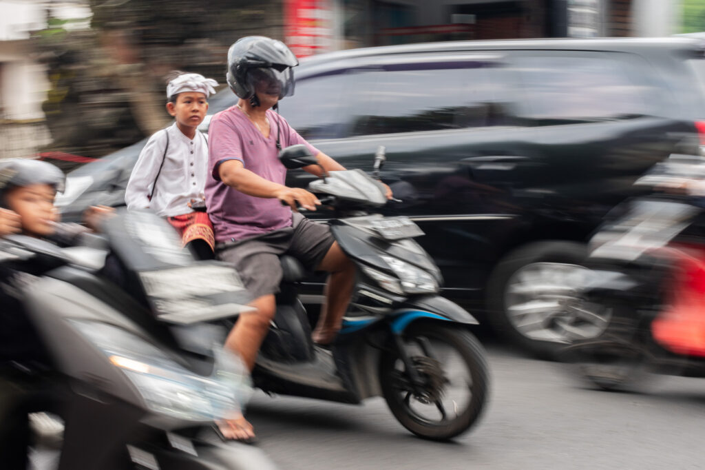 Un homme portant un casque et un t-shirt violet roule en scooter, avec un garçon en costume traditionnel assis derrière lui. Ils se frayent un chemin dans la circulation, entourés de voitures et d'autres scooters dont les contours sont flous.
