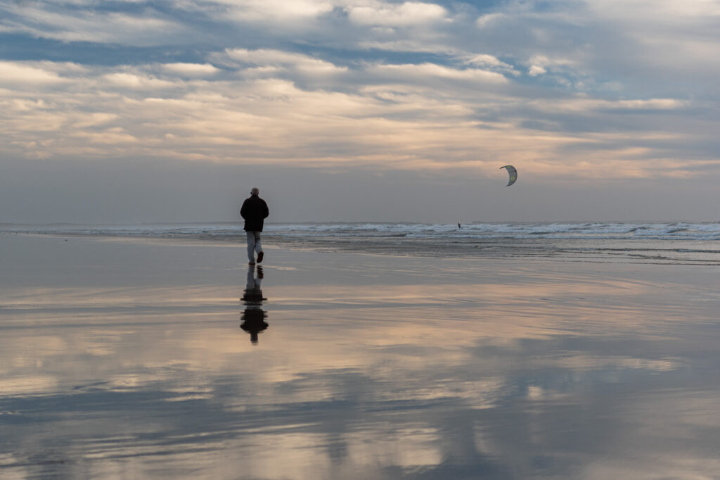 Une personne marche seule sur une plage mouillée au coucher du soleil, tandis que des nuages spectaculaires et le ciel se reflètent dans le sable. On aperçoit au loin, près des vagues, un kitesurfeur.