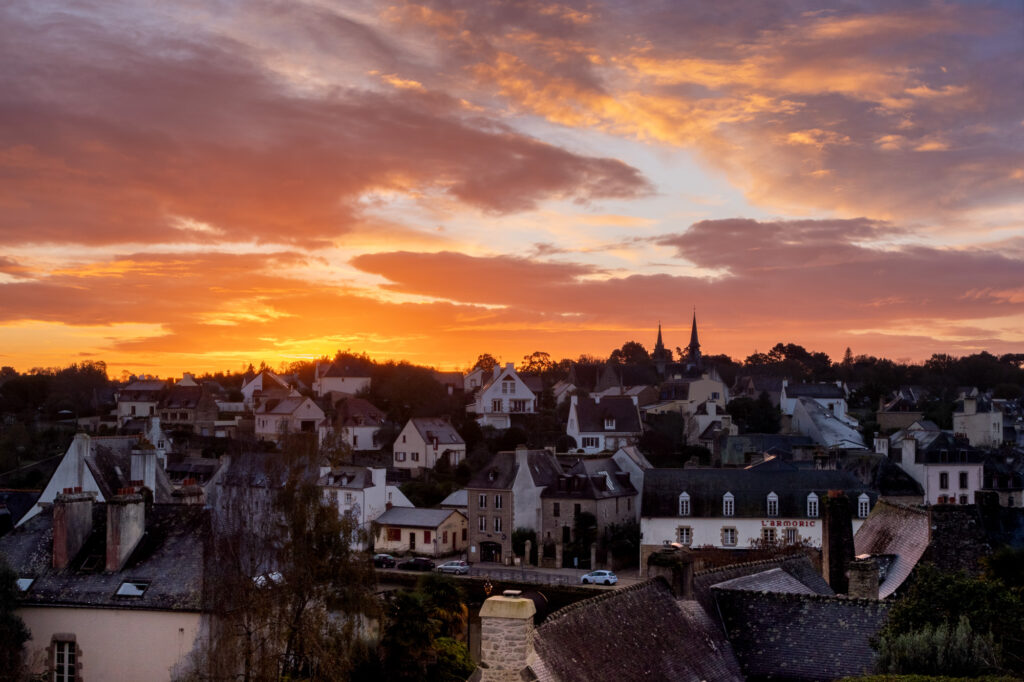 Un village pittoresque, avec ses charmantes maisons et le clocher de son église, se détache sur un ciel de coucher de soleil aux teintes spectaculaires d'orange et de violet, créant ainsi une scène nocturne paisible.