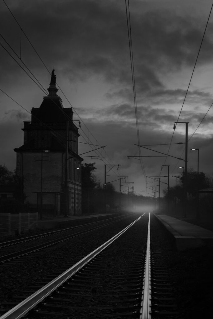 À la tombée de la nuit, les voies ferrées s'étendent à perte de vue, reflétant la lumière sous un ciel nuageux. Un vieux bâtiment doté d'une tour se dresse à gauche, tandis que des lampadaires bordent le quai désert à droite.