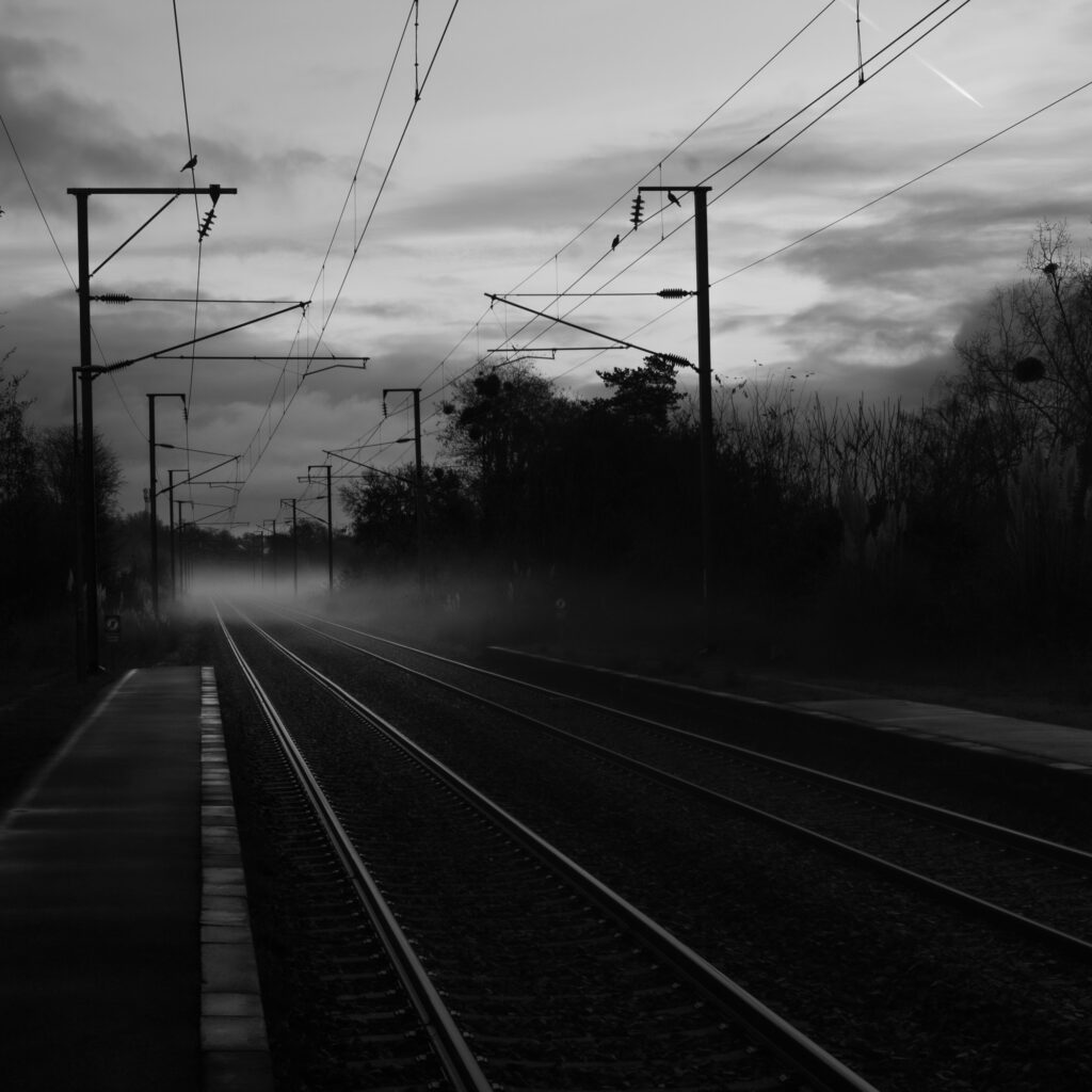 À la tombée de la nuit, les voies ferrées, enveloppées de brume, s'étendent à perte de vue, flanquées de lignes électriques et d'arbres se découpant sur le ciel nuageux, où l'on aperçoit une légère traînée de condensation.
