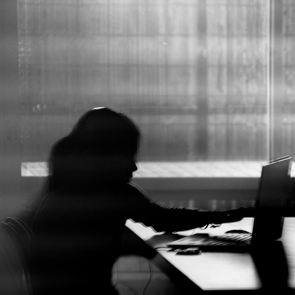 La silhouette d'une personne assise à un bureau, devant un ordinateur portable, avec une lumière douce filtrant par une fenêtre en arrière-plan, créant une atmosphère mélancolique et contemplative.