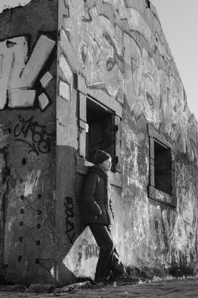 Par une journée ensoleillée, une personne portant un bonnet et une veste est adossée au mur d'un immeuble délabré et couvert de graffitis. L'immeuble est doté de grandes fenêtres et des ombres se dessinent sur sa façade.