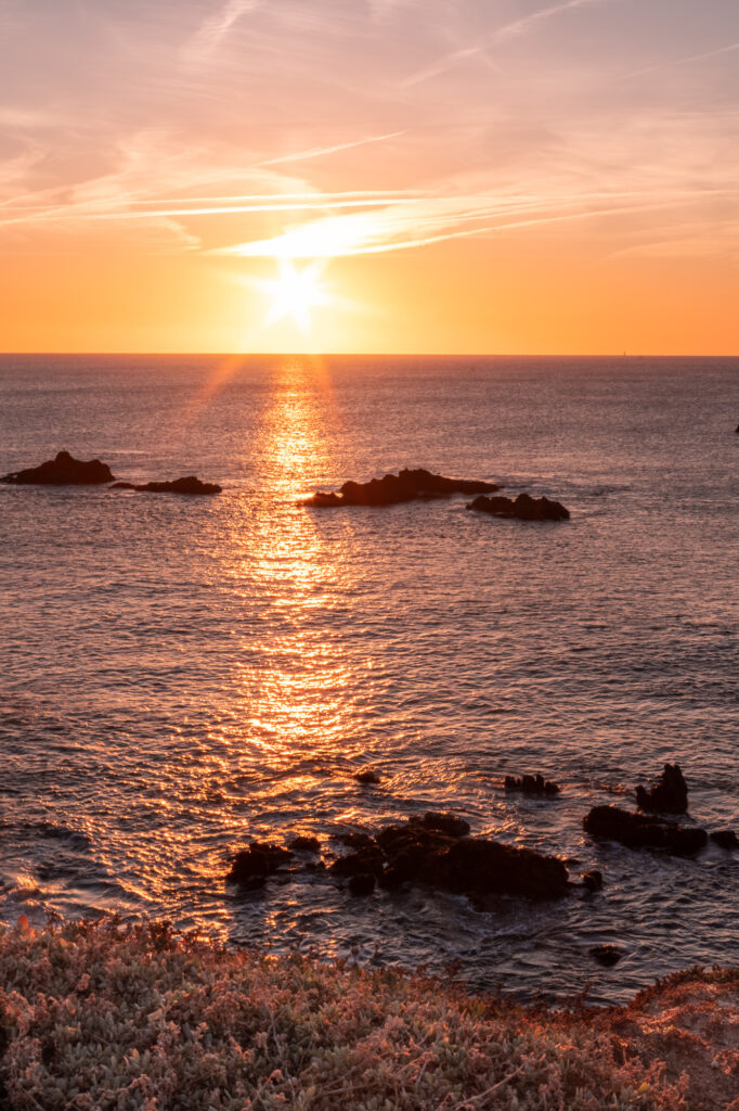 Coucher de soleil sur l'océan : le soleil, bas à l'horizon, projette un reflet doré sur l'eau. On distingue des rochers sombres et des vagues douces, avec au premier plan une étendue herbeuse et un ciel strié de nuages orange et roses.