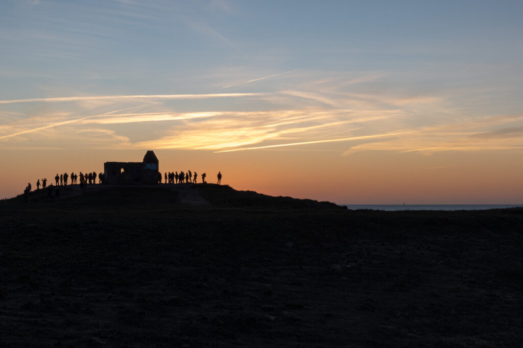 Silhouettes de personnes debout sur une colline au coucher du soleil, rassemblées près d'un petit bâtiment, avec en arrière-plan un ciel coloré et des traînées de nuages.