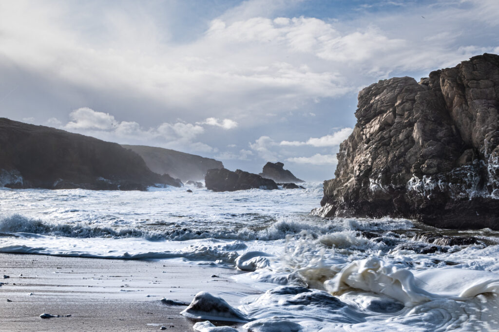 Les vagues viennent s'écraser sur une plage de sable bordée de part et d'autre par de grandes falaises rocheuses, sous un ciel partiellement nuageux. L'eau écumeuse tourbillonne au ras du rivage et les rayons du soleil filtrent à travers les nuages.