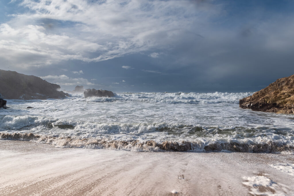 Les vagues viennent s'écraser sur une plage de sable bordée de falaises rocheuses, sous un ciel bleu nuageux ; on distingue au premier plan de l'écume blanche et des remous.