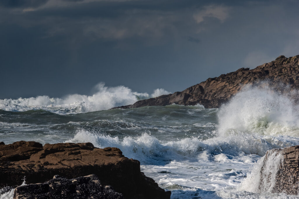 Les vagues viennent se briser contre les falaises rocheuses et les rochers sombres sous un ciel nuageux et maussade, projetant dans les airs des gerbes d'eau et de l'écume blanche. La mer agitée semble puissante et spectaculaire.