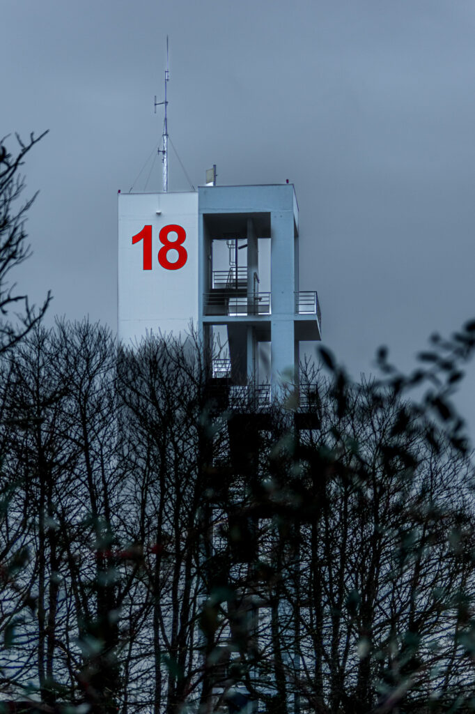 Une haute tour blanche, sur laquelle est inscrit en gros un « 18 » rouge, se détache au-dessus des arbres dénudés, sur fond de ciel gris et nuageux.