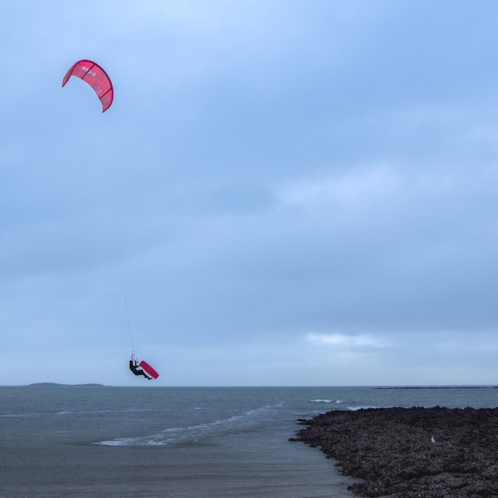 Une personne pratique le kitesurf bien au-dessus de l'eau, accrochée à un cerf-volant rouge. La mer est calme et le ciel est couvert ; on aperçoit en arrière-plan un littoral rocheux et des îles au loin.