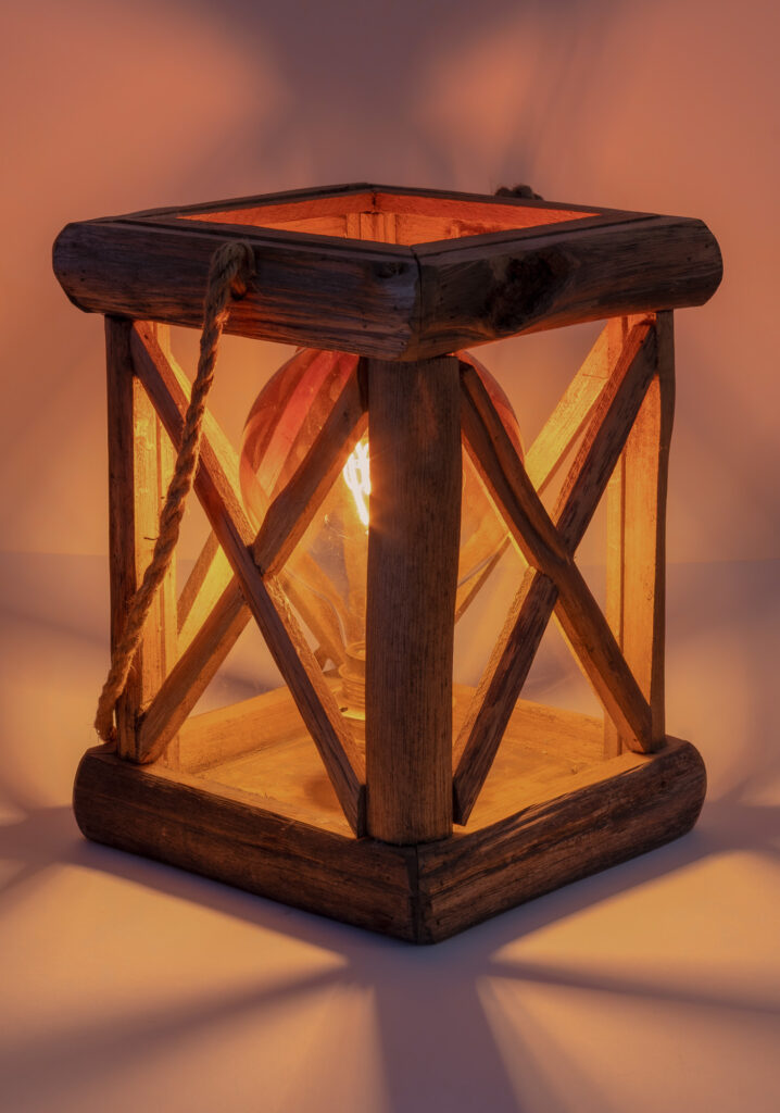 Une lanterne rustique en bois, dotée d'une poignée en corde et de parois en verre, éclairée de l'intérieur par une ampoule à la lueur chaleureuse qui projette de douces ombres sur la surface environnante.