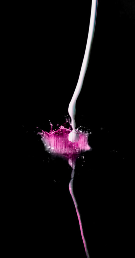 Un jet de liquide blanc éclabousse une petite mare de liquide rose sur fond noir, créant des ondulations et des gouttelettes dont le reflet est saisissant.