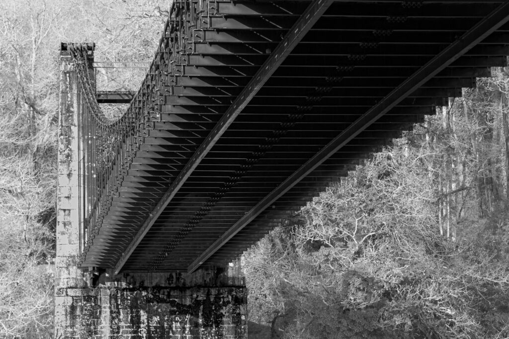 Photo en noir et blanc montrant la face inférieure d'un pont suspendu aux poutres métalliques, enjambant une rivière, avec en arrière-plan des arbres dépouillés de leurs feuilles.