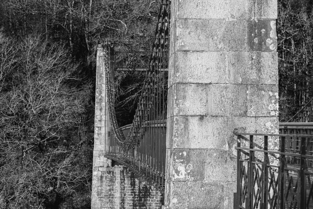 Photo en noir et blanc d'un pont suspendu, avec un grand pilier en pierre au premier plan et des arbres dépouillés à l'arrière-plan. Le pont est muni de garde-corps métalliques et de câbles.
