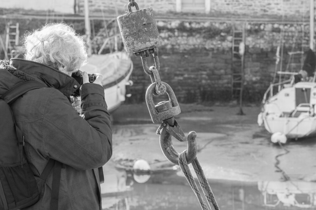 Une personne aux cheveux bouclés, portant un sac à dos, prend une photo près d'un port, en cadrant un grand crochet d'amarrage métallique au premier plan ; on aperçoit des bateaux et un mur de pierre à l'arrière-plan. L'image est en noir et blanc.