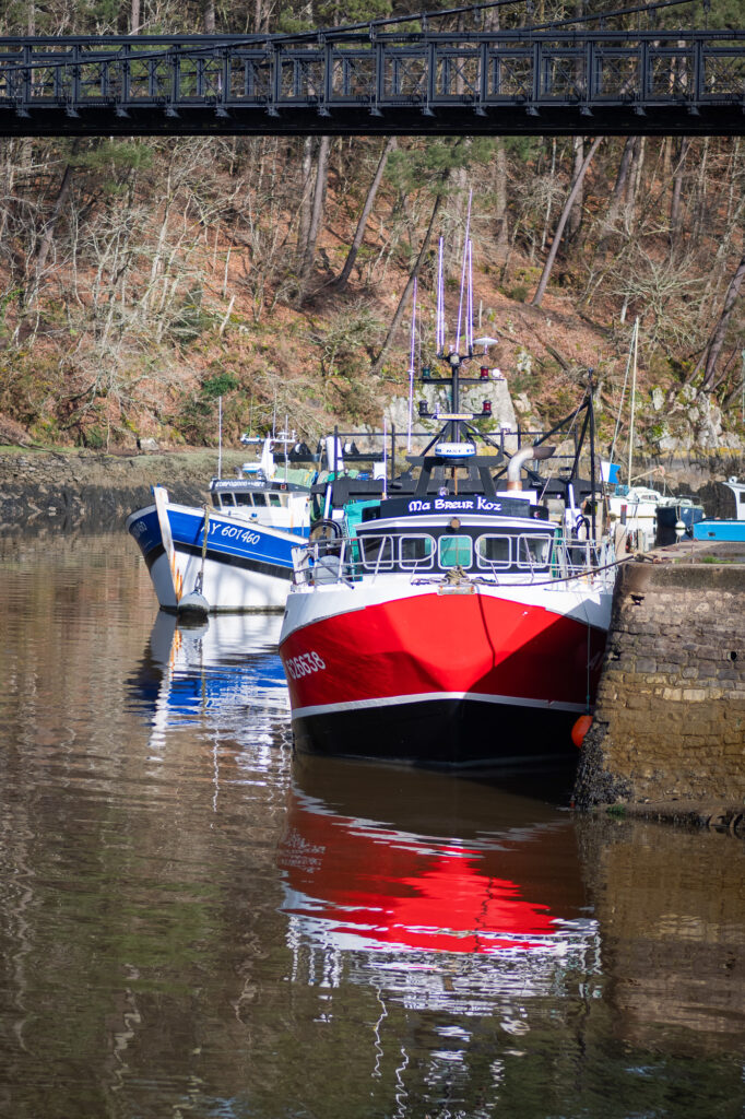 Trois bateaux de pêche, dont un rouge et blanc baptisé « Ma Broere Koz », sont amarrés le long d'un mur de pierre dans une rivière calme, sous un pont en fer noir, avec des arbres et de la végétation en arrière-plan.