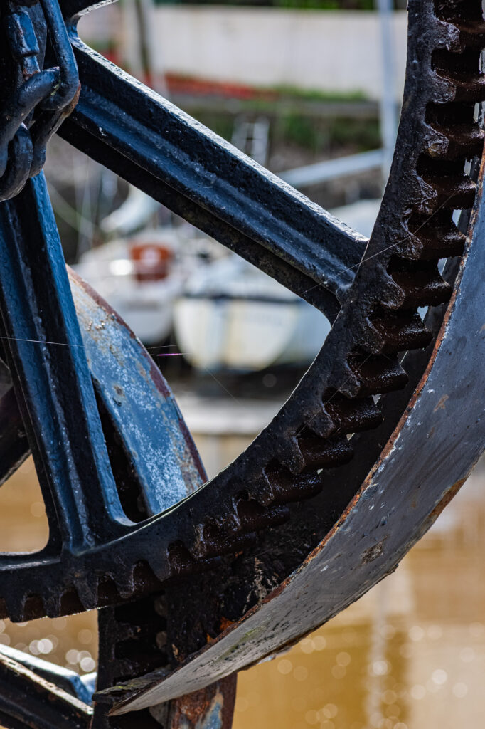 Gros plan sur un grand engrenage métallique bleu, patiné par les intempéries, dont les dents sont visibles, placé en diagonale. À l'arrière-plan flou, on aperçoit des bateaux et de l'eau, ce qui suggère un décor de quai ou de port de plaisance.