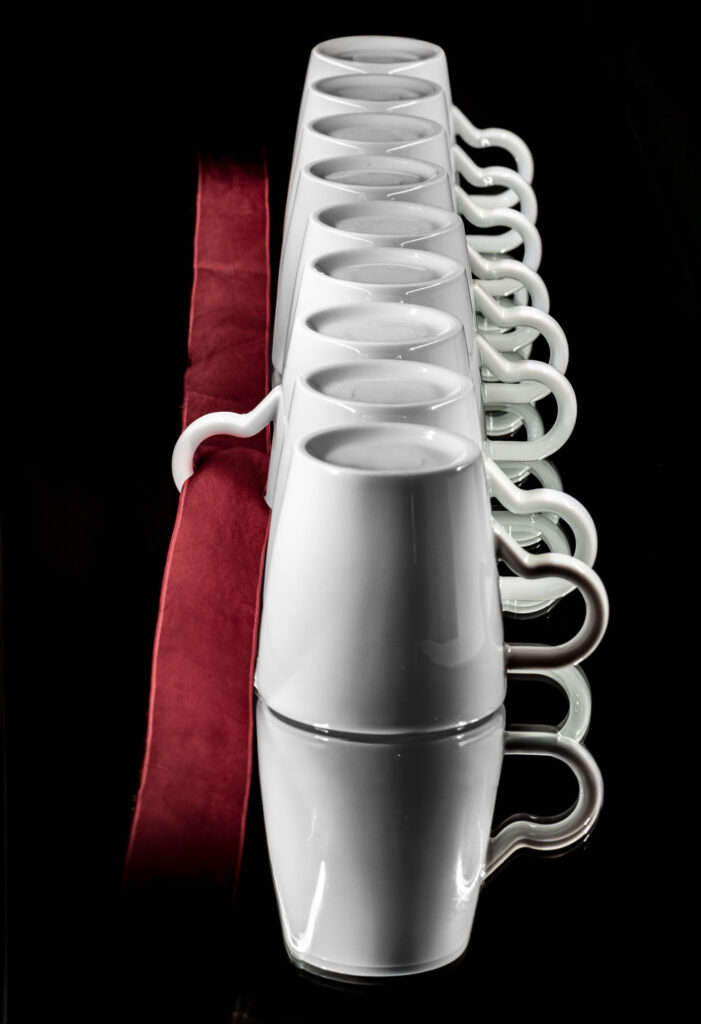 Huit tasses en céramique blanche sont disposées à l'envers en une rangée bien ordonnée à côté d'un tissu rouge plié, le tout sur fond noir. Les anses des tasses forment un motif répétitif sur le côté.