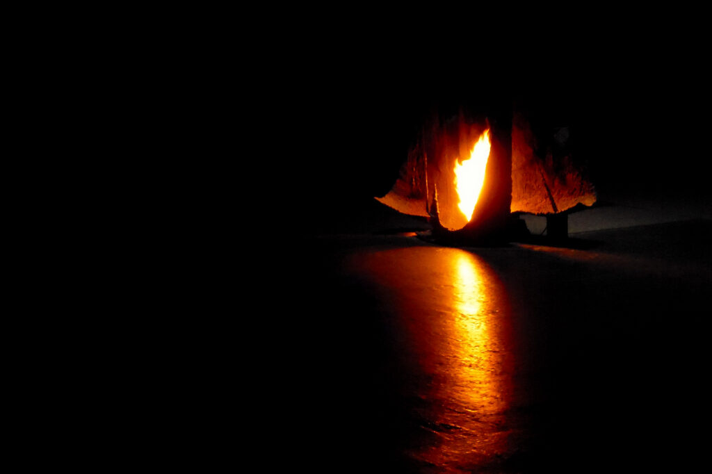 Un petit feu vif brûle à l'intérieur d'une structure en forme de tente, projetant une lueur orange et chaleureuse ainsi que des reflets sur un sol sombre et vide, entouré d'ombres profondes.