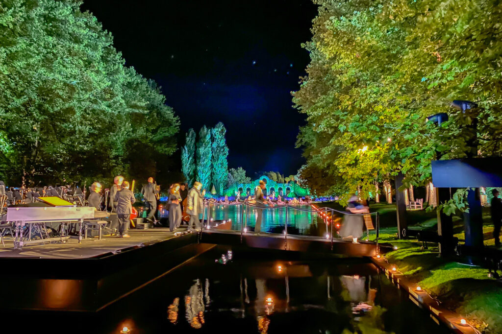 Une scène de concert nocturne animée en plein air au bord d'un canal, avec des musiciens et des spectateurs sur scène, des lumières colorées illuminant les arbres et des reflets scintillants sur l'eau.