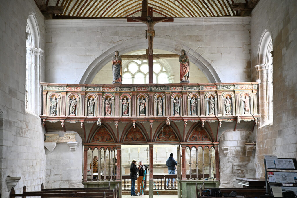 L'intérieur d'une église en pierre, avec un jubé sculpté et peint représentant des figures religieuses, un grand crucifix au-dessus, des bancs en bois en dessous, et plusieurs personnes debout au fond de la nef.