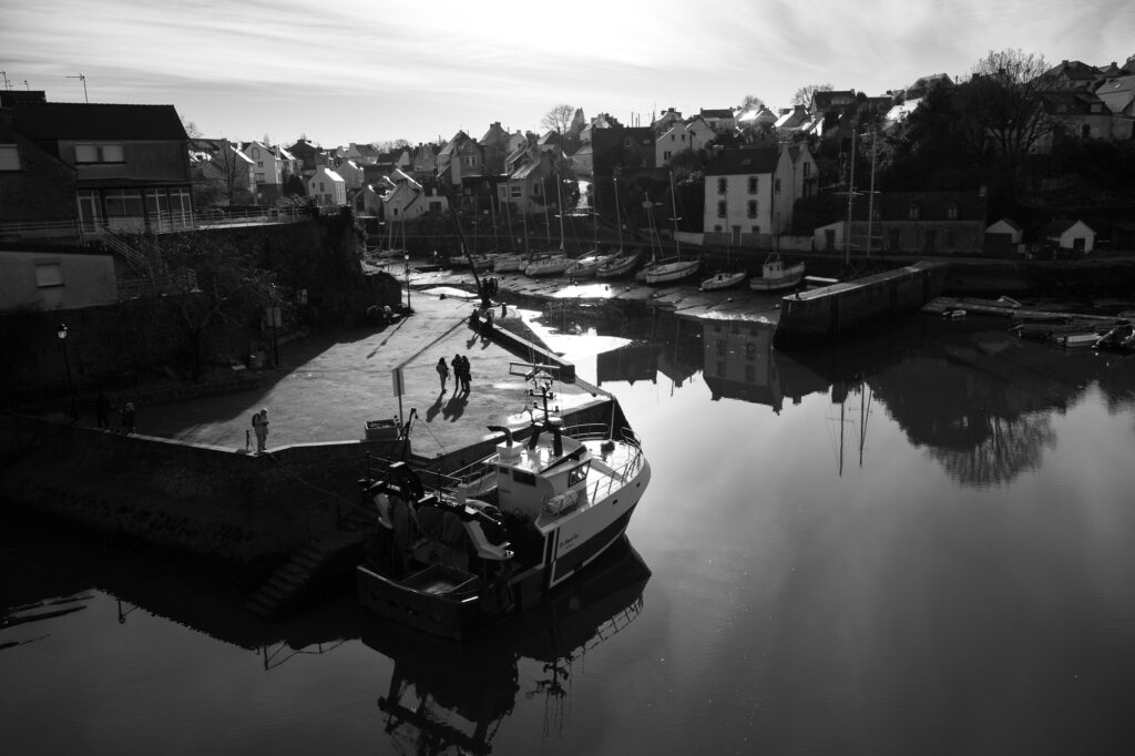 Une photo en noir et blanc d'un port paisible où est amarré un bateau, avec des eaux calmes reflétant le ciel, quelques personnes se promenant sur le quai et des maisons bordant le front de mer sous un ciel partiellement nuageux.